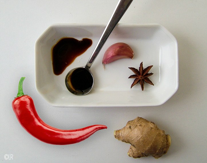 Een paar van de ingrediënten om zelf ketjap te maken. Lepeltje ketjap manis met een teen roze knoflook, een steranijs, stukje gemberwortel en pepertje