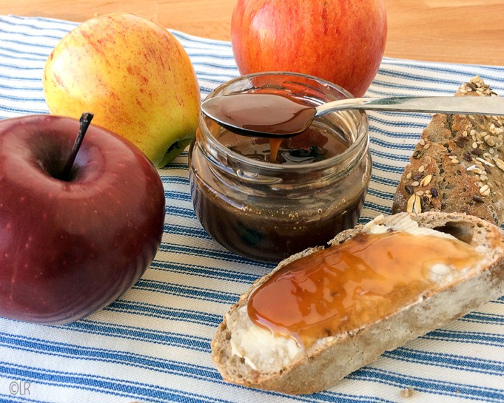 Temidden van wat appels staat een potje met licht vloeibare appelstroop die van de lepel loopt. Er naast een snee volkorenbrood met pitjes besmeerd met roomboter en appelstroop