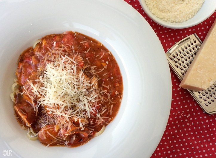 Bord spaghetti met saus en geraspte Parmezaanse kaas. Er naast een stuk Parmezaanse kaas op een rasp en geraspte kaas in een schaaltje.