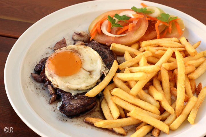 Bord met een mooi gebakken biefstuk en een gebakken ei er bovenop. Geserveerd met friet en een eenvoudige tomatensalade.