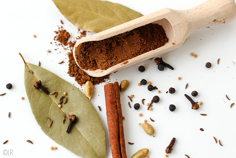 Een schepeltje geroosterde kruiden omringd door laurier, kaneel, kardemom, kruidnagel, mosterd en peper.