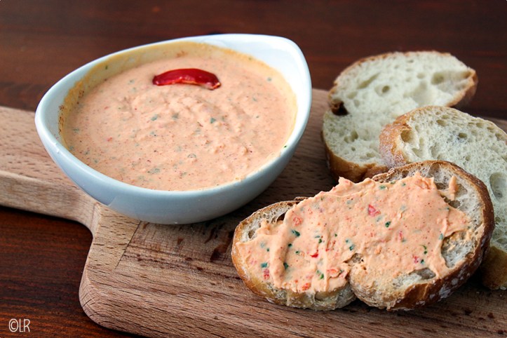 Schaaltje kruidenboter met erop een schijfje paprika. Ernaast wat stokbrood besmeerd met deze boter.