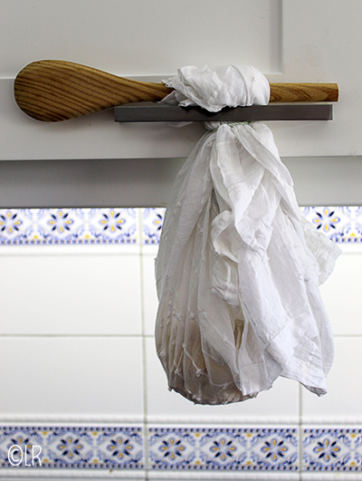 Een kaasdoek gevuld met kwark die met een houten lepel is opgehangen aan het handvat van een keukenkastje. 